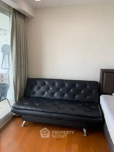 Modern living room with sleek black leather sofa and wooden flooring, adjacent to a balcony with outdoor seating.
