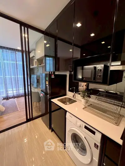 Modern kitchen with sleek black cabinets, washing machine, and microwave, adjacent to a stylish living area.