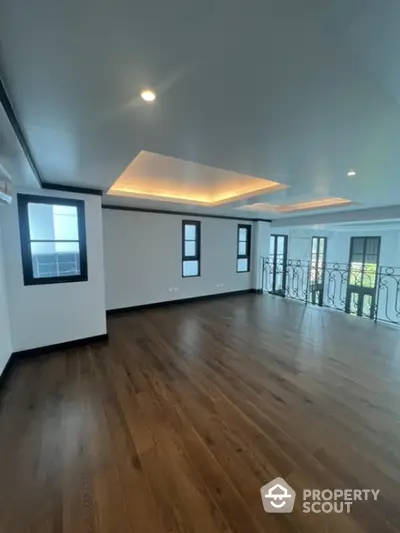 Spacious and elegant living room with polished hardwood floors, sophisticated tray ceiling design, and abundant natural light from multiple windows leading to a charming balcony.