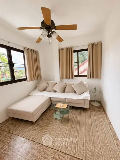 Cozy living room with natural light, featuring a beige sofa, glass tables, and ceiling fan.