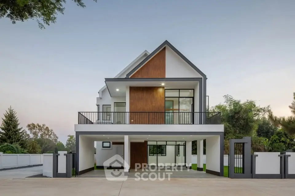 Modern two-story house with sleek design and spacious balcony, surrounded by lush greenery and a paved driveway.