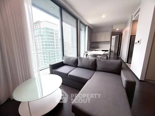 Modern living room with large windows and city view, featuring a sleek gray sofa and glass coffee table.