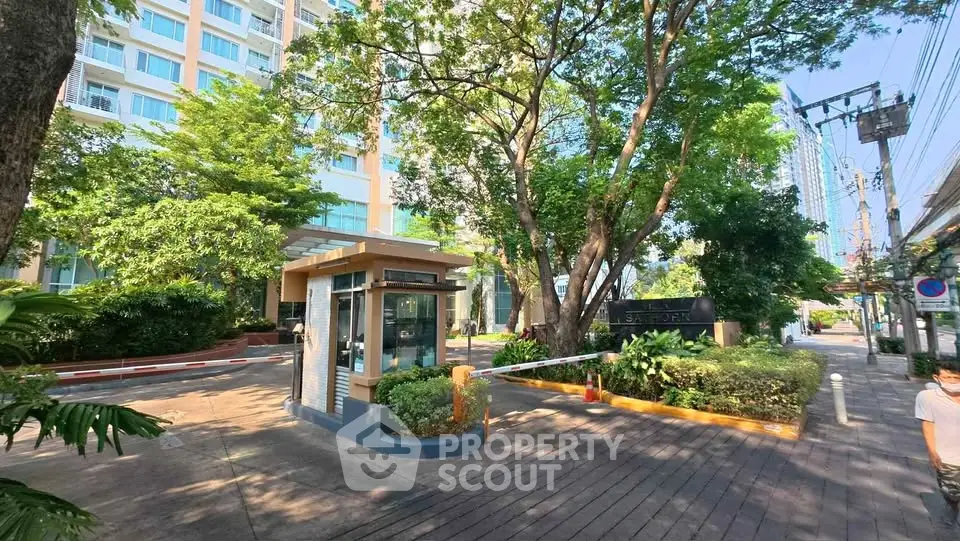 Modern condominium entrance with lush greenery and security booth in urban setting.