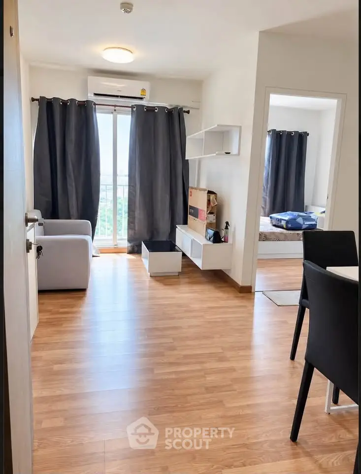 Modern apartment interior with open living room and bedroom, featuring wooden flooring and large windows.