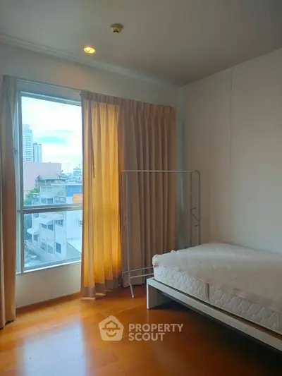 Bright bedroom with large window and city view, featuring wooden flooring and minimalist decor.