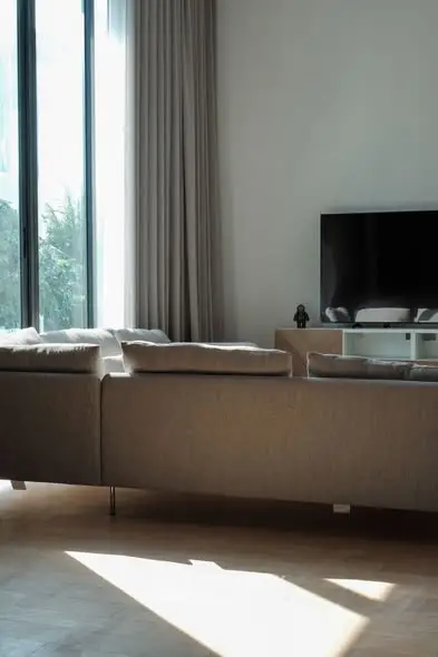 Modern living room with large sofa and flat-screen TV, featuring floor-to-ceiling windows.