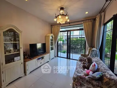 Charming living room with elegant chandelier and garden view, featuring cozy floral furniture and modern entertainment setup.