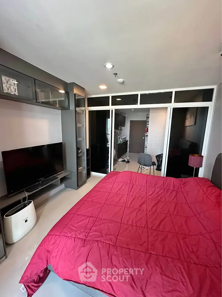 Modern bedroom with red bedding, TV, and glass partition leading to kitchen.