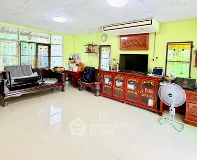 Spacious living room with traditional wooden furniture and bright green walls, featuring large windows and air conditioning.