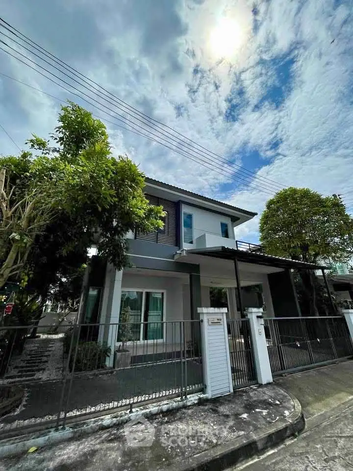 Modern two-story house with gated entrance and lush greenery under a bright sky.