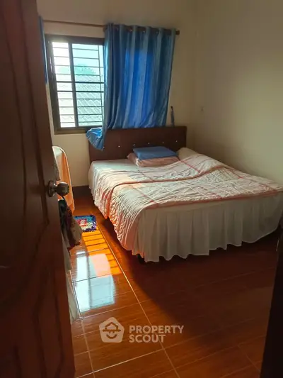 Cozy bedroom with natural light and wooden flooring, featuring a comfortable bed and blue curtains.