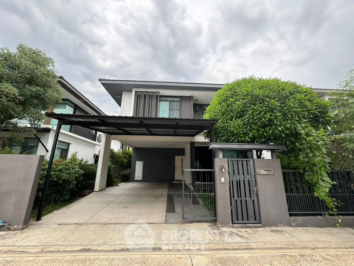 Modern two-story house with carport and lush greenery under a cloudy sky.