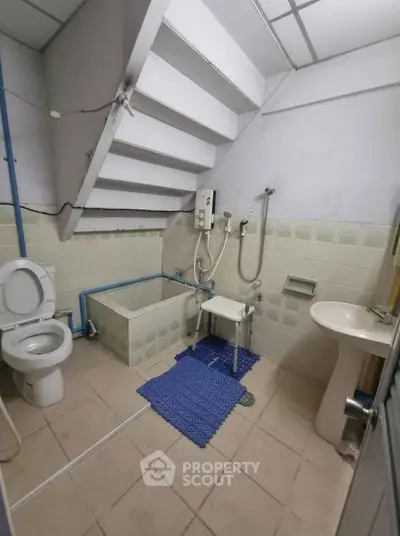 Compact bathroom with unique staircase overhead, featuring toilet, sink, and shower area.