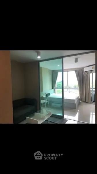 Modern apartment with glass partition separating living room and bedroom, featuring sleek furniture and natural light.