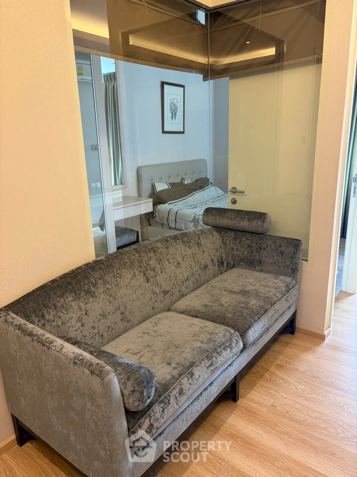 Modern living room with plush gray sofa and glass partition overlooking a cozy bedroom.