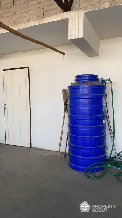 Spacious utility area with large blue water tank and broom against white wall.