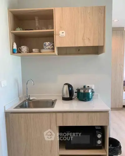 Compact kitchen with wooden cabinets, sink, microwave, and kettle in modern apartment.