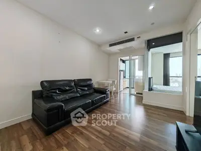 Spacious modern living room with sleek black leather sofa and wooden flooring, leading to a bright bedroom.