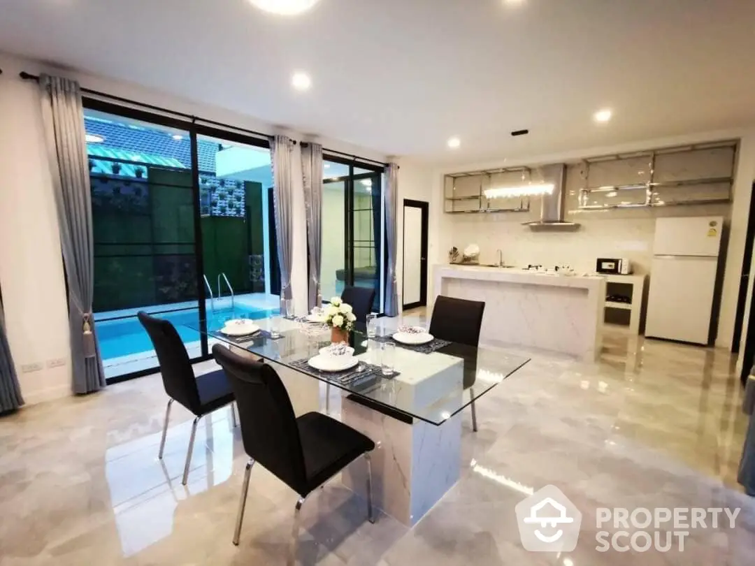 Spacious and modern open-plan living area with sleek dining set, leading to a well-equipped kitchen and view of the pool through large glass doors.