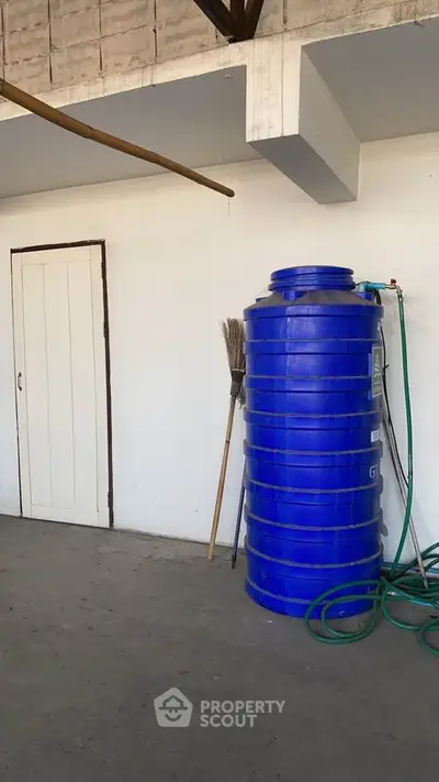 Spacious utility area with large blue water tank and broom against white wall.