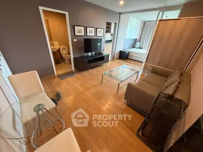 Modern living room with wooden flooring, glass table, and cozy seating area leading to a bedroom.