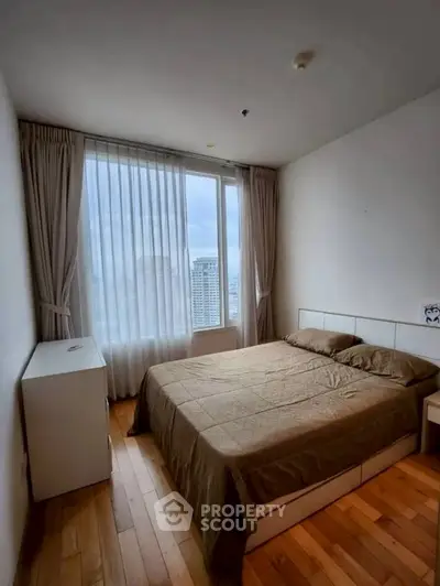 Cozy bedroom with city view and natural light, featuring wooden flooring and modern furnishings.