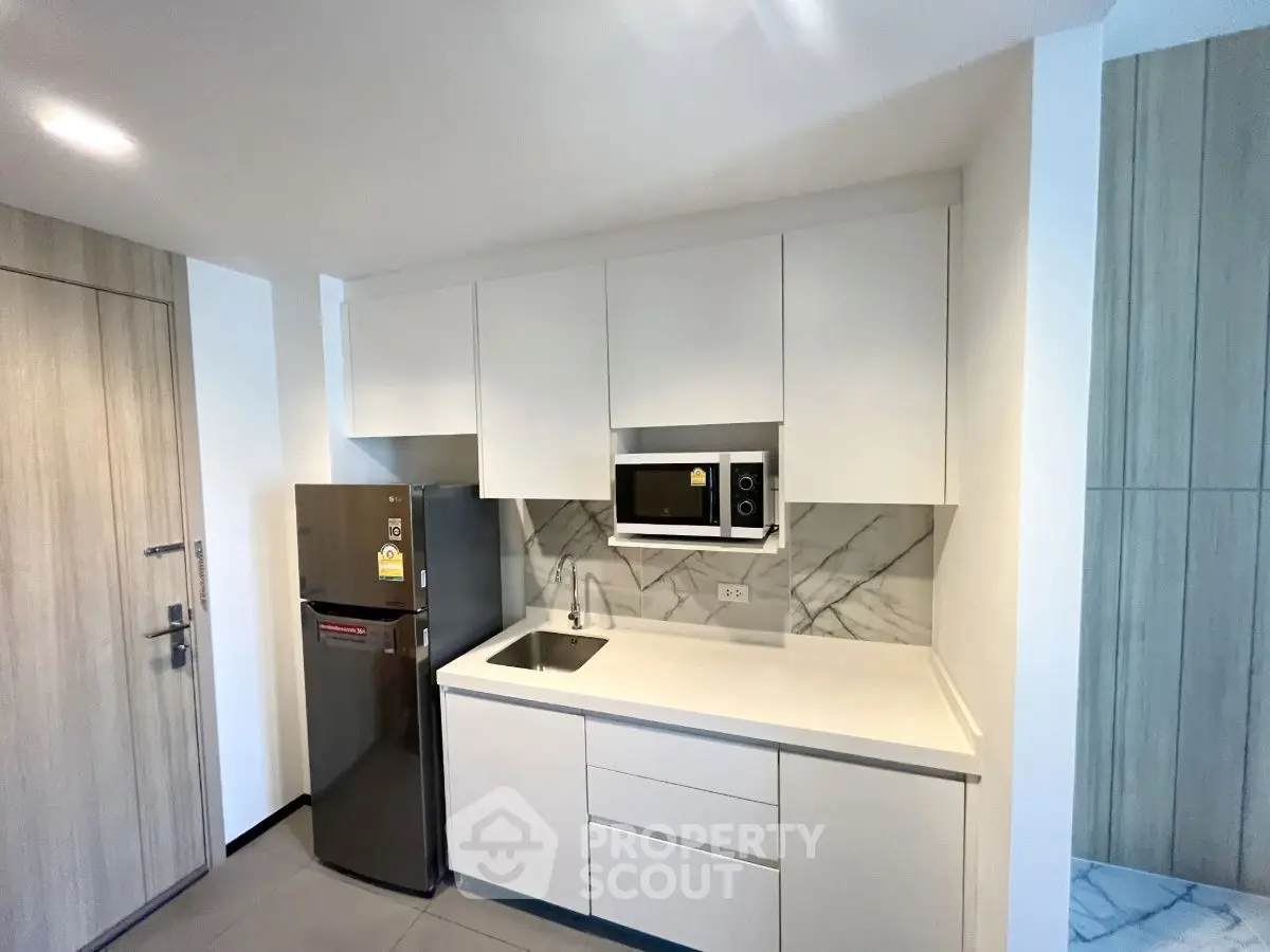 Modern kitchen with sleek white cabinets and built-in microwave, featuring a compact fridge and elegant marble backsplash.