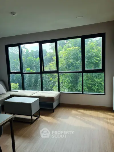 Bright living room with large windows and lush green view, featuring modern furniture and wooden flooring.