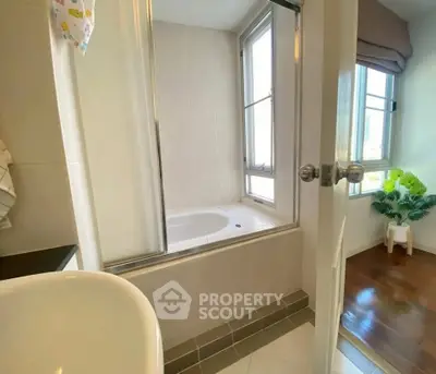 Spacious bathroom with bathtub and natural light, adjacent to a bright room with wooden flooring.