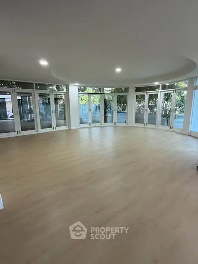 Spacious circular living room with large windows and wooden flooring, offering a serene garden view.