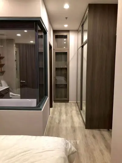 Modern bedroom with glass-enclosed bathroom and wooden flooring, featuring sleek storage solutions.
