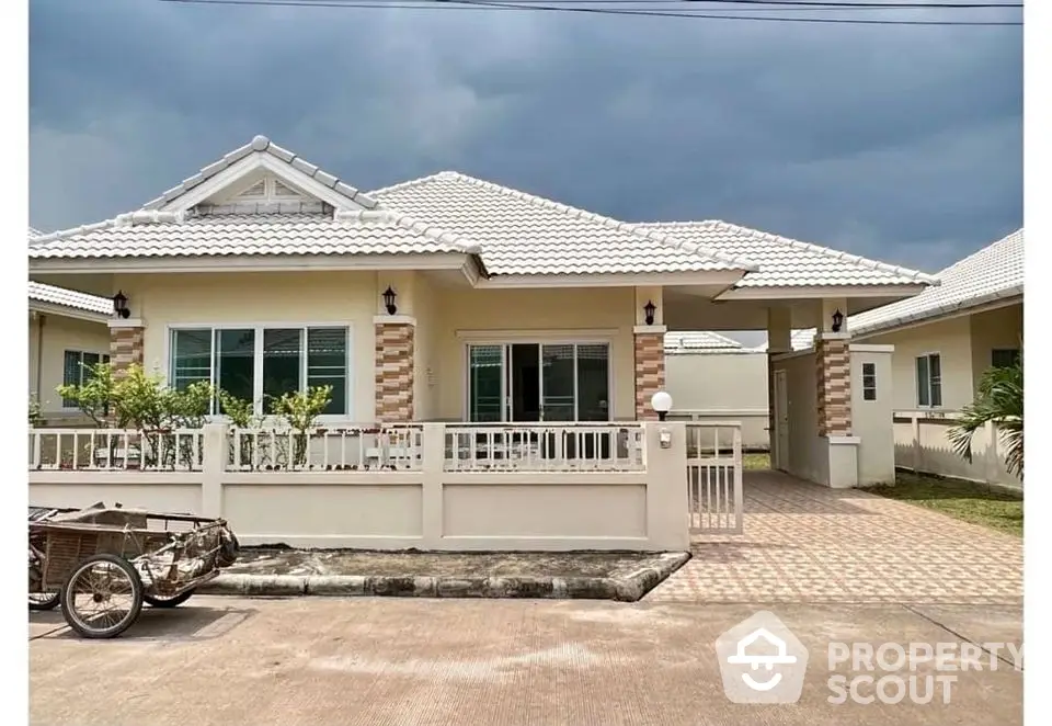 Charming single-story house with white tile roof and spacious driveway under cloudy sky.