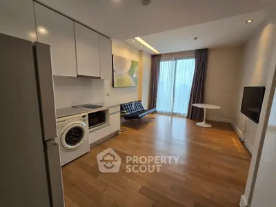 Modern living room with integrated kitchen and washing machine, featuring sleek furniture and large window.