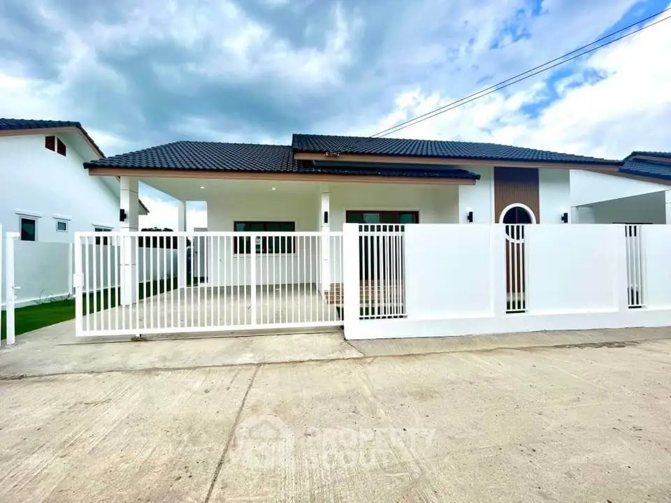 Modern single-story house with white fence and spacious driveway under cloudy sky.