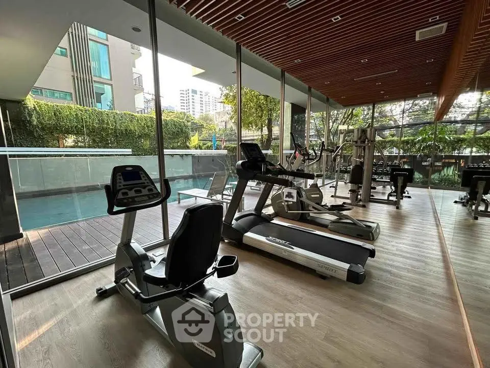 Modern gym with glass walls overlooking pool and greenery, featuring state-of-the-art fitness equipment.