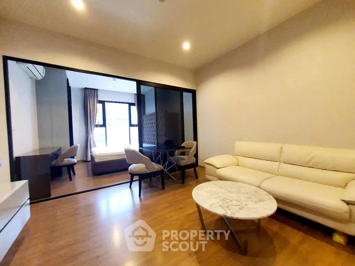 Modern living room with elegant furniture and wooden flooring, featuring a view into a cozy bedroom.
