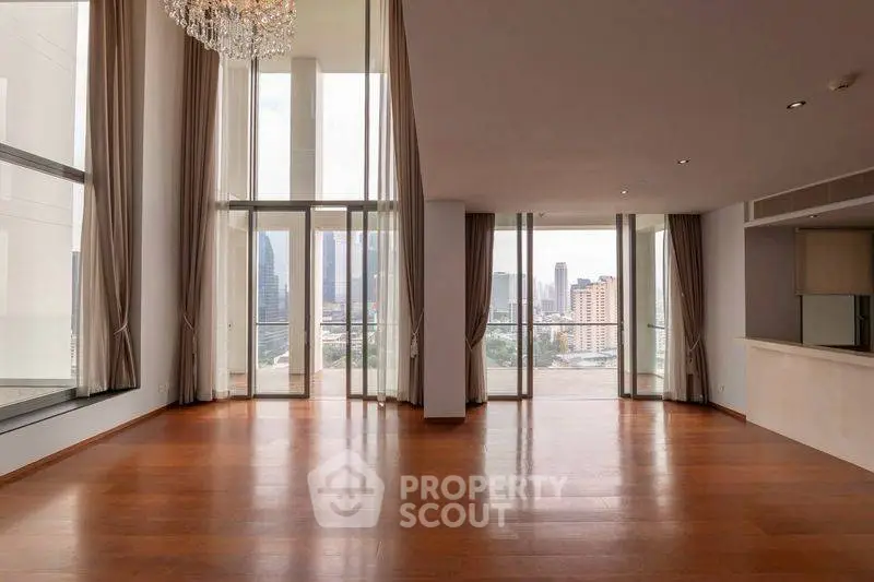 Spacious modern living room with floor-to-ceiling windows and city view, featuring elegant chandelier and wooden flooring.