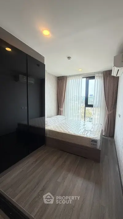 Modern bedroom with large window and city view, featuring sleek black wardrobe and wood flooring.