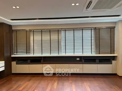 Modern living room with wooden flooring and large windows with blinds, featuring built-in storage units.