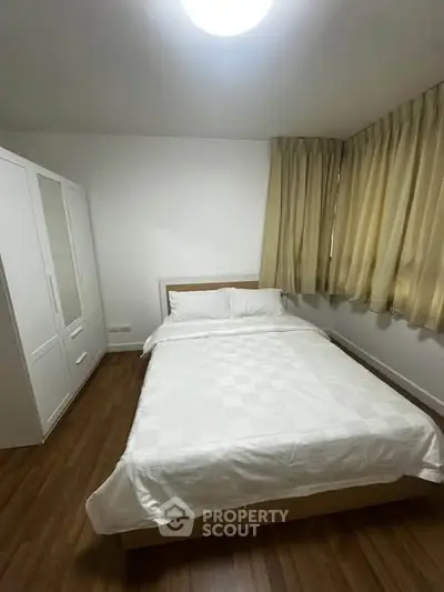Cozy bedroom with wooden flooring, white wardrobe, and large window with curtains.