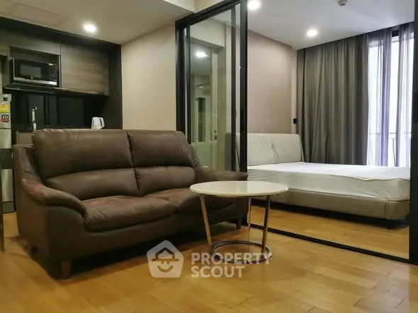 Modern studio apartment with cozy living area and bedroom, featuring sleek glass partition and wooden flooring.