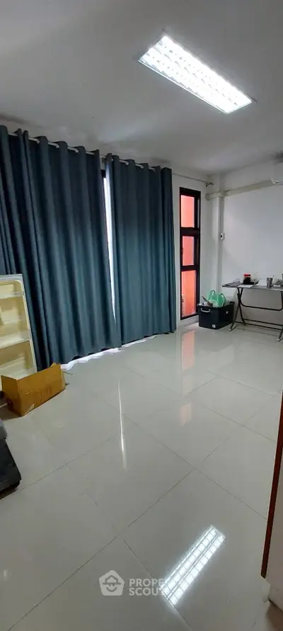 Spacious and well-lit living space with glossy tiled flooring, large windows draped with elegant teal curtains, and a modern ceiling light fixture, perfect for a comfortable urban lifestyle.