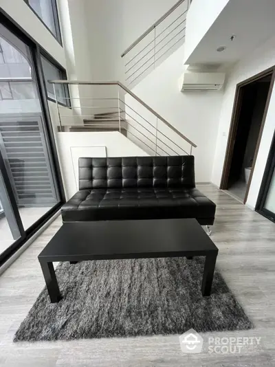 Modern living room with sleek black sofa and coffee table, featuring large windows and stylish staircase.