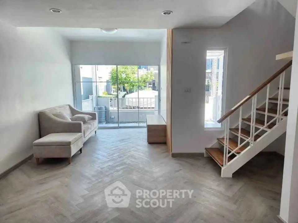 Modern living room with sleek staircase and large window offering natural light and outdoor view.