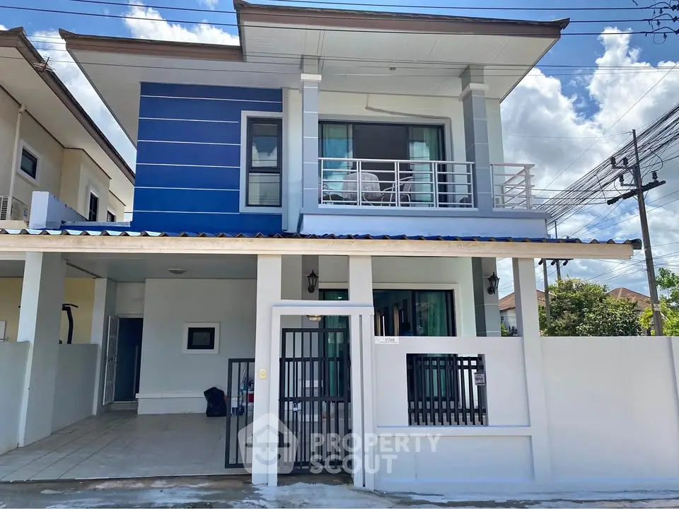 Modern two-story house with blue accents and a balcony, featuring a gated entrance and carport.