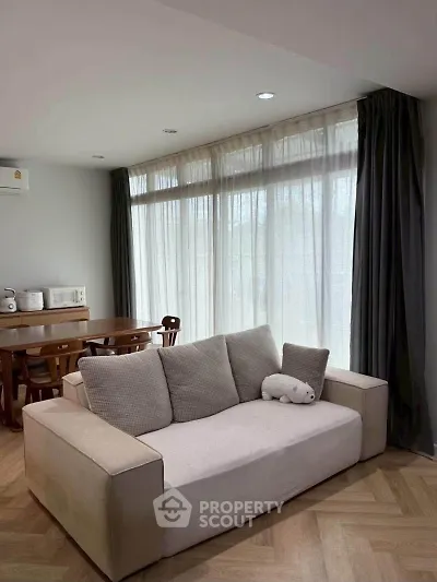 Cozy living room with modern sofa and dining area, featuring large windows and elegant curtains.