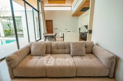 Luxurious open-plan living room with modern kitchen and pool view, featuring plush sofa and high ceilings.