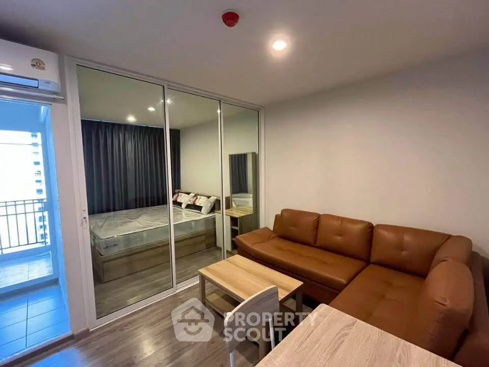 Modern living room with brown leather sofa and glass partition leading to bedroom, featuring sleek wooden flooring.