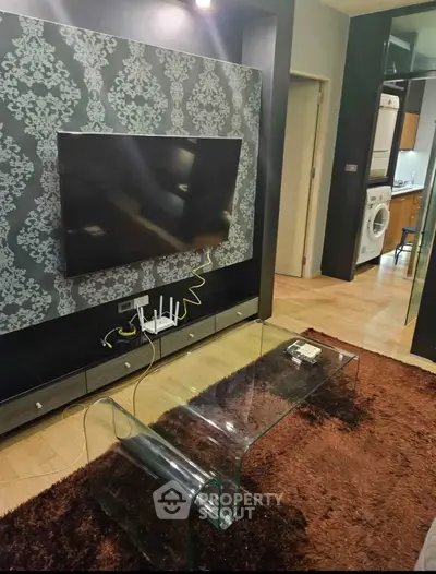 Modern living room with stylish wallpaper, glass coffee table, and wall-mounted TV, leading to a kitchen with appliances.