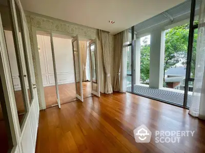 Spacious room with wooden flooring and large windows opening to a balcony with lush greenery view.